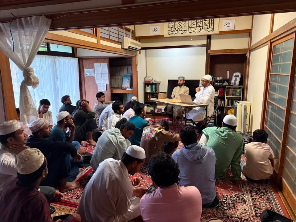 A group of men wearing kufi caps sit on a carpeted floor in a converted Japanese-style room, listening to two speakers seated at a table in front of a bookshelf and Arabic calligraphy on the wall.