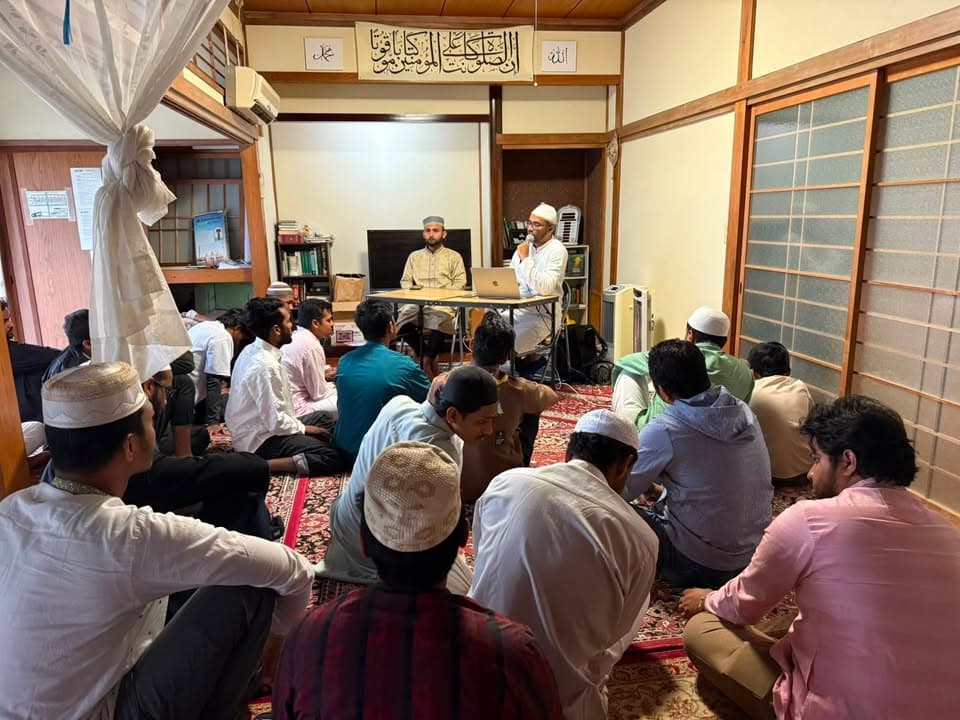 A group of men in traditional caps and clothing sit cross-legged on patterned rugs in a Japanese-style room, listening to two speakers seated at a table with a laptop at the front, beneath Arabic calligraphy on the wall.