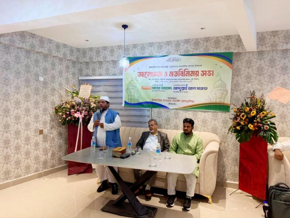 A man in a white robe and blue vest stands speaking into a microphone beside a table, while two other men sit on a sofa listening, in front of a banner with Bengali text and floral arrangements on either side.
