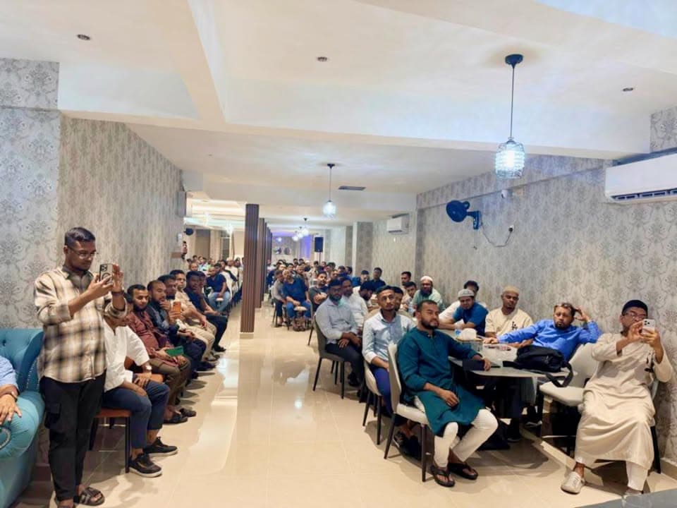 A long hall filled with rows of seated men listening attentively, some using their phones to take photos, while a few stand near the front recording video.