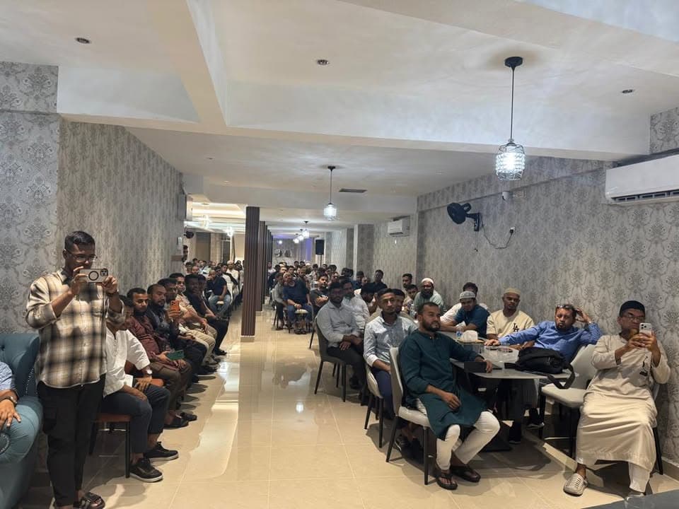 A long, narrow hall filled with rows of men seated on chairs, some in traditional attire, listening attentively while a man near the front records the scene on his phone.