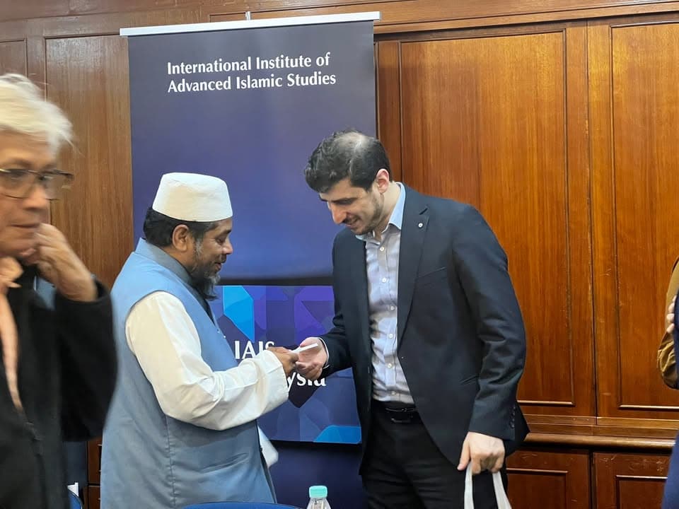 Two men exchange a business card while standing in front of a banner reading "International Institute of Advanced Islamic Studies," inside a wood-paneled room with other attendees nearby.
