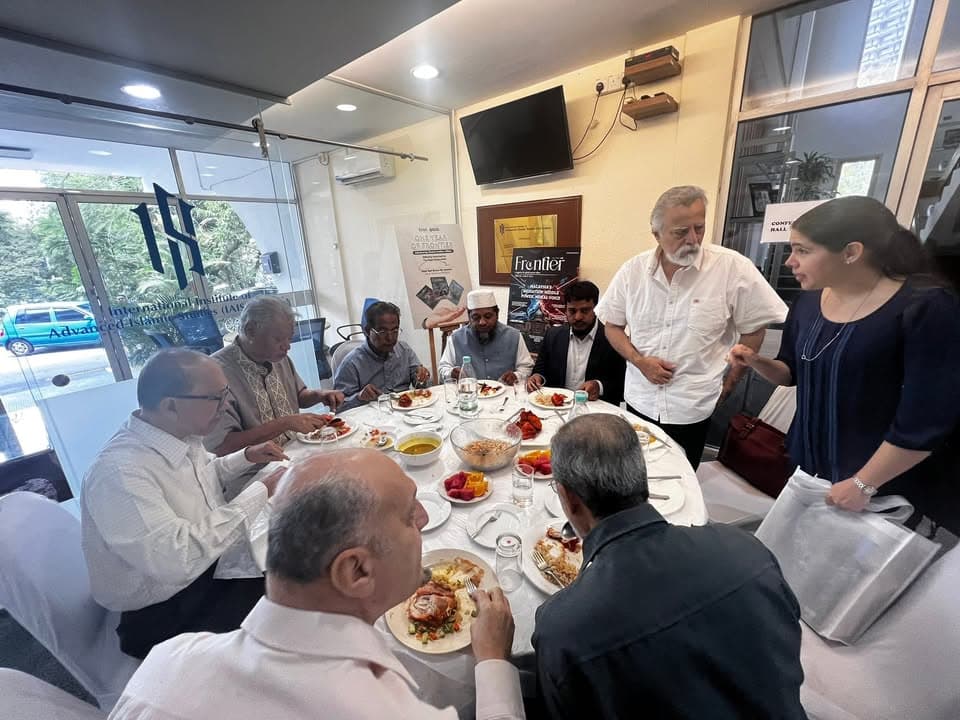 A group of men and women, some wearing formal shirts and one in a white cap and vest, sit and stand around a round table with plates of food at the International Institute of Advanced Islamic Studies (IAIS) office.