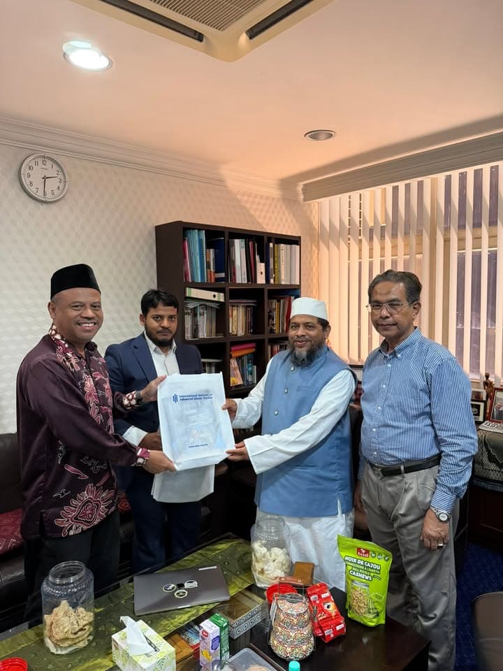 Four men stand together in an office, with two of them jointly holding a folder printed with the "International Institute of Advanced Islamic Studies" logo, in front of a bookshelf and a table with snacks.
