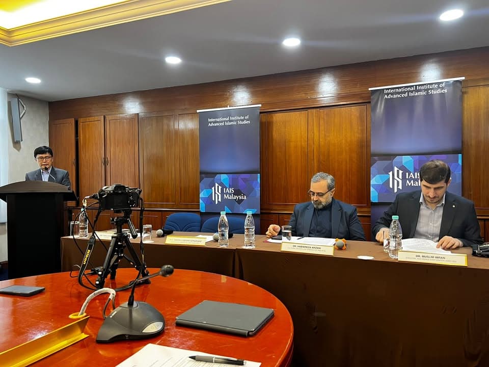 A man speaks at a podium while two seated panelists at a conference table listen, in front of banners reading "International Institute of Advanced Islamic Studies" and "IAIS Malaysia" in a wood-paneled hall.