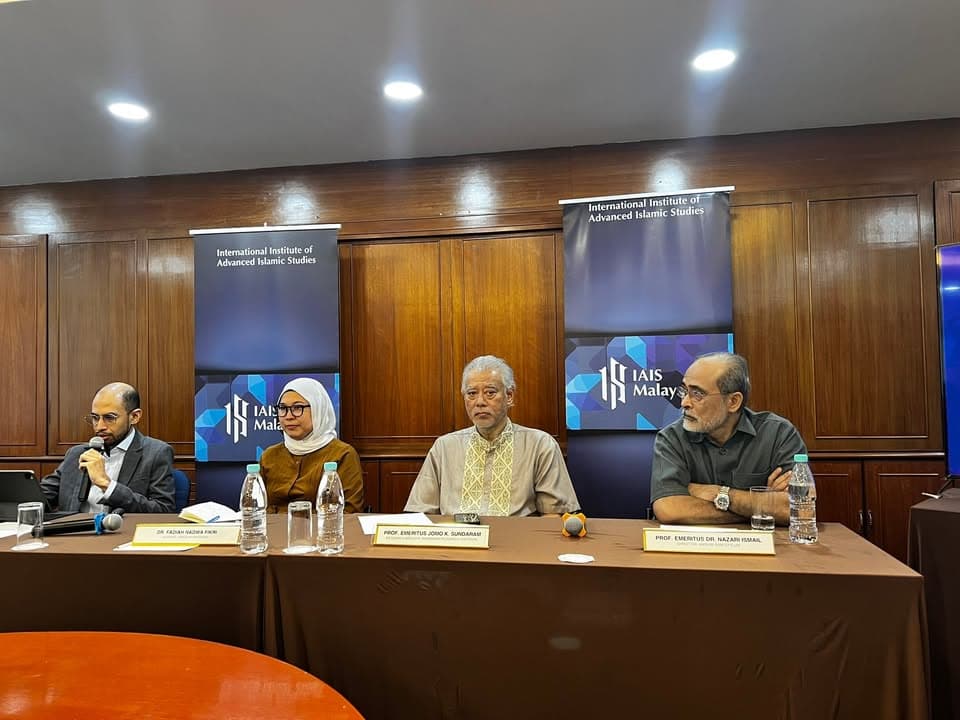 Four panelists sit behind a long table with nameplates and water bottles, in front of two banner stands reading "International Institute of Advanced Islamic Studies," with one panelist speaking into a microphone.