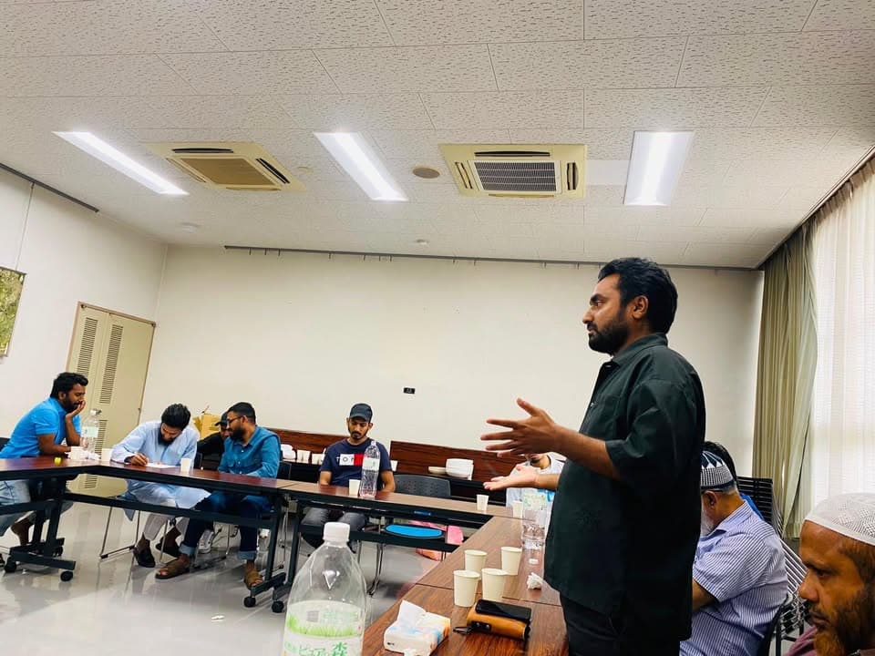A man standing in a meeting room gestures with both hands while speaking to a group of seated men at tables with water bottles and paper cups, some in traditional attire and caps.