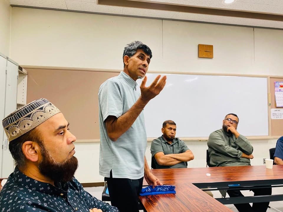 A man stands and gestures while speaking to a seated group of men in a meeting room with a whiteboard and clock on the wall behind him.
