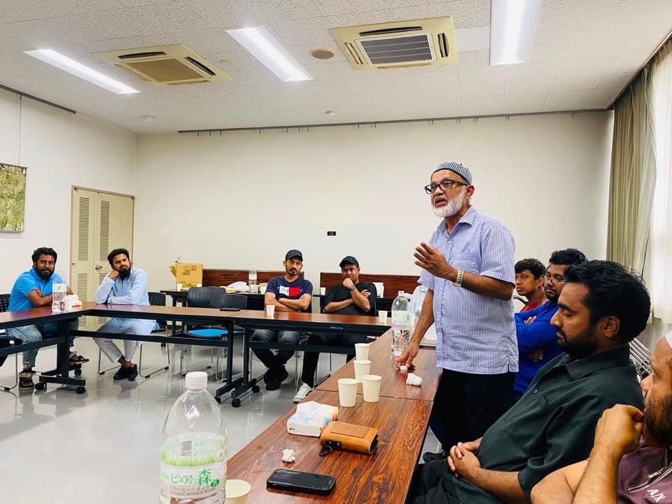 An older man wearing a striped shirt and prayer cap stands and speaks with his hand raised while a group of seated men listen around tables set with water bottles and paper cups in a meeting room.