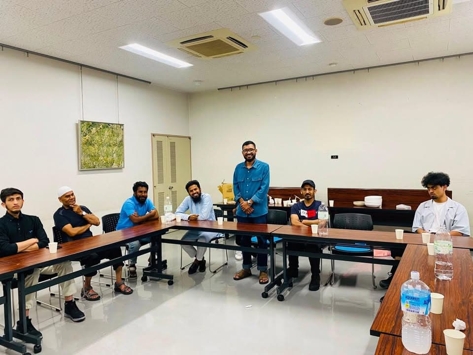 A group of men sit and stand around tables arranged in a U-shape in a community room, smiling toward the camera, with water bottles and paper cups on the tables.