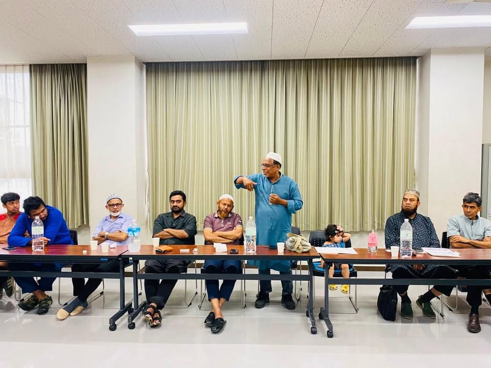 A man in a light blue kurta and cap stands and speaks while gesturing, in front of a row of seated men and a young child at long tables in a community hall with beige curtains.