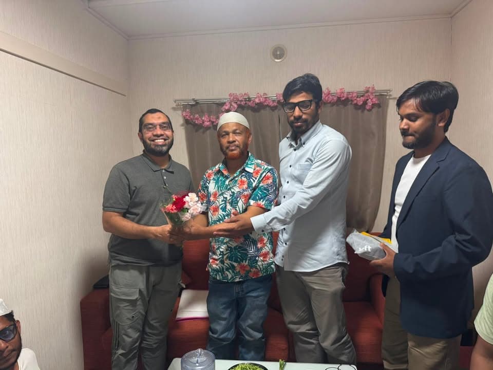 Four men stand indoors in a small room, with one presenting a bouquet of flowers to another as the group smiles for the photo.