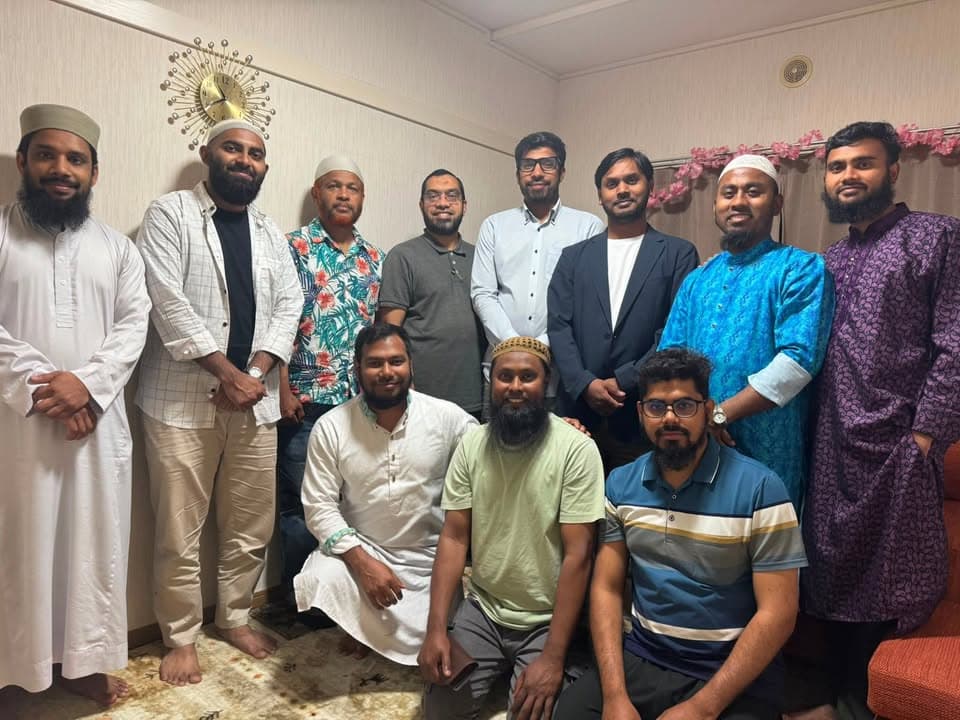 A group of eleven men, some standing and some kneeling, pose together for a group photo indoors, several wearing traditional kufi caps and long tunics.