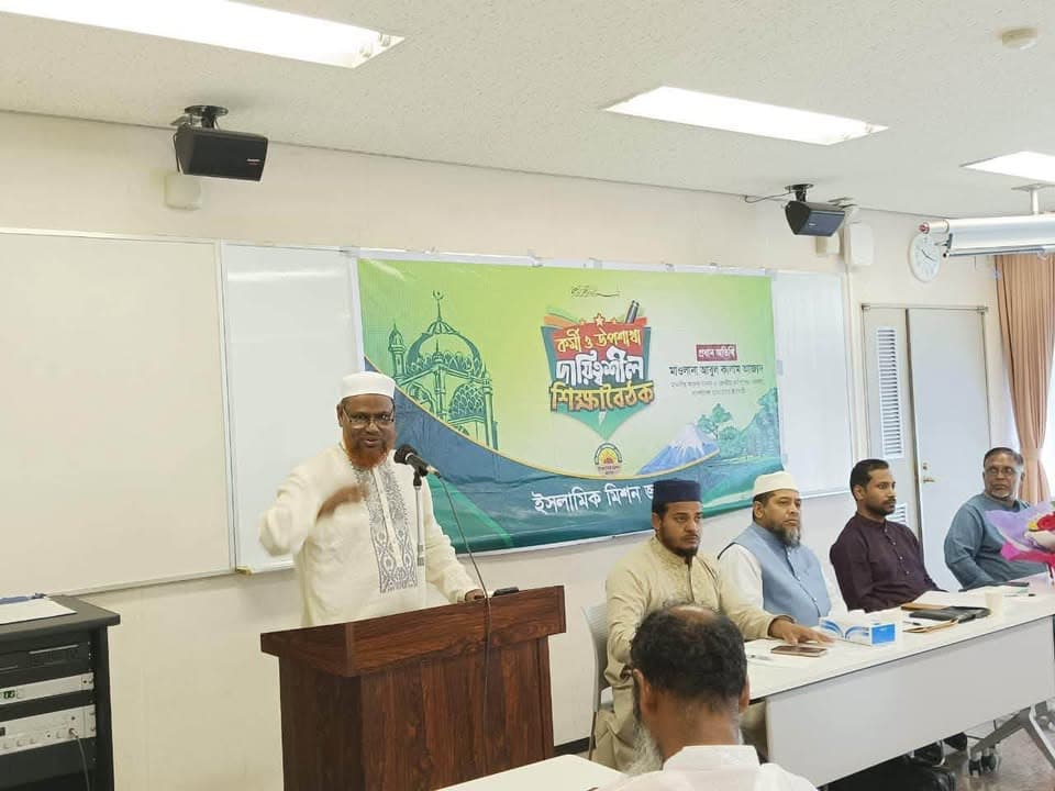 A man in white traditional attire speaks at a podium microphone in front of a green banner with Bengali text, while several other men sit at tables listening in a meeting room with a whiteboard and projector screen in the background.
