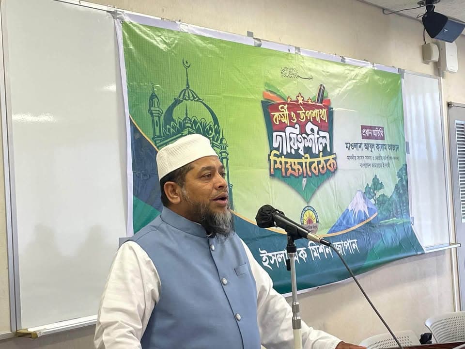 A man wearing a white cap and light blue vest speaks into a microphone at a podium, in front of a large green banner with Bengali text and a mosque illustration, next to a whiteboard.