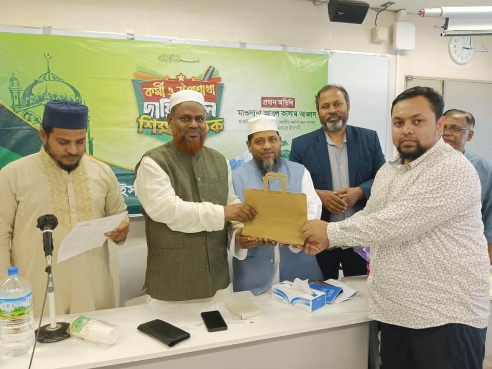 Several men stand in front of a green banner with Bengali text as one man hands a paper gift bag to another man in a patterned shirt, while others look on in what appears to be a meeting room.