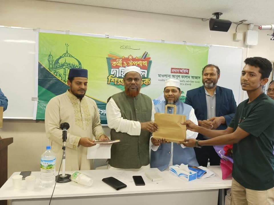A group of men stand behind a table on a stage, several wearing white caps and traditional attire, as one man in a green vest hands a brown envelope to a younger man in a green polo shirt, in front of a green banner with Bengali text and a mosque logo.