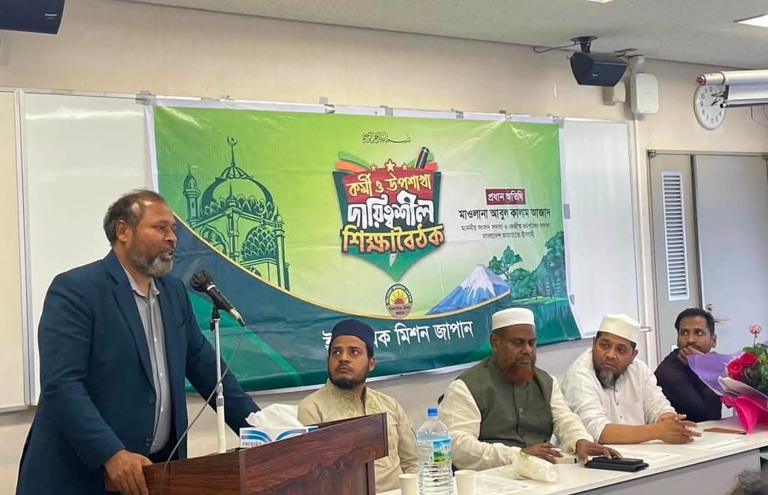 A man in a dark blazer speaks at a podium with a microphone, while four seated men in Islamic caps listen at a table in front of a green banner with Bengali text and mosque graphics, in a classroom-style meeting room.