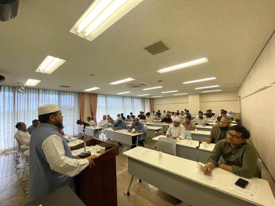 A speaker wearing a white cap stands at a podium addressing rows of men seated at tables with notebooks and papers in a fluorescent-lit conference room.
