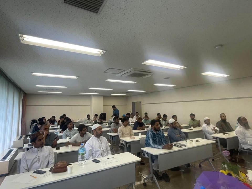 Rows of men seated at white desks in a classroom-style hall, listening attentively during a training session.