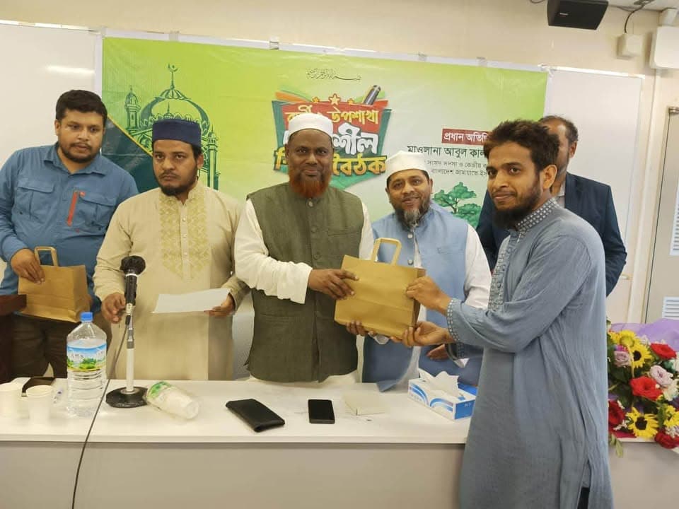 A group of men stand behind a table at an indoor event, with one man handing a paper gift bag to another man in front of a green banner with Bengali text.