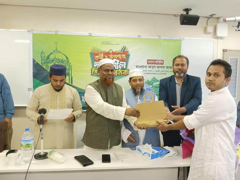 A man in a green vest hands a wrapped package to another man in white at the front of a meeting room, with several other men standing behind them beside a green banner with Bengali text and a table holding microphones and papers.