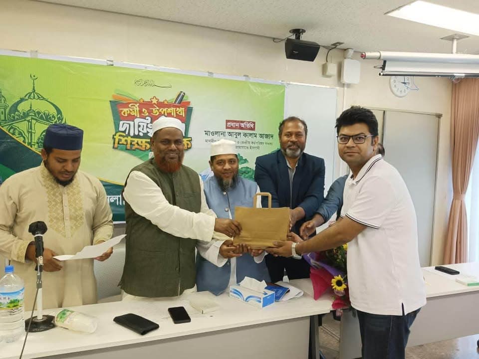 A group of men stand behind a table in an indoor meeting room, presenting a paper gift bag and a bouquet of flowers to a man in a white polo shirt, in front of a green banner with Bengali text.