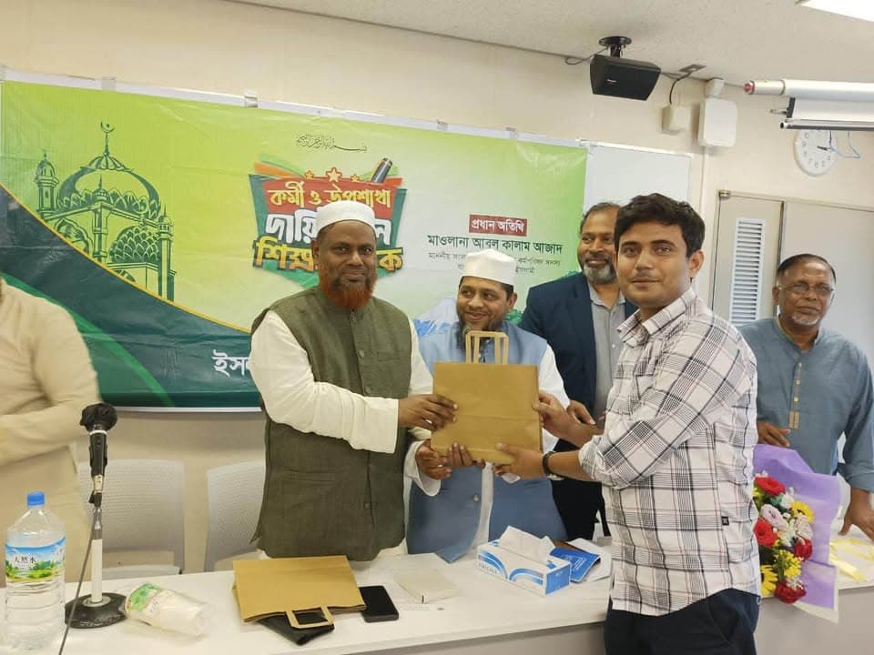 Men stand at the front of a meeting room in front of a green banner with Bengali text, presenting a paper gift bag to a participant while others look on.