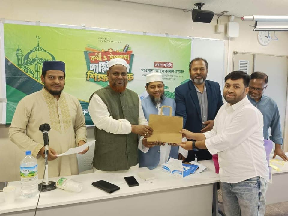 Five men stand behind a table in front of a green banner with Bengali text, with two men in the center presenting a paper gift bag to each other as a group photo moment during an indoor event.