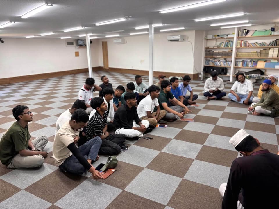 A group of young men sit cross-legged in rows on a checkered-tile floor inside a mosque hall, listening attentively, with bookshelves and a scholar in traditional attire visible at the front.