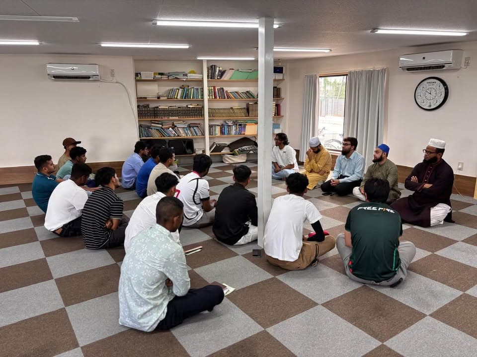 A group of men sit cross-legged in a circle on a checkered floor in a mosque room lined with bookshelves, listening to several elder men seated together speaking to them.