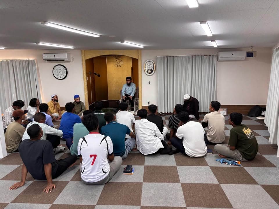 A group of men and young students sit cross-legged on a checkered carpet inside a mosque prayer hall, listening to a speaker seated near the mihrab.