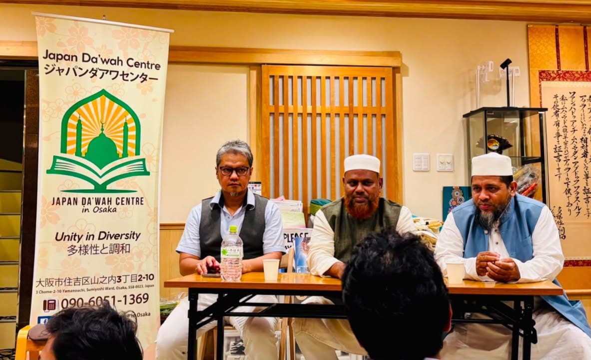 Three men sit behind a table facing an audience inside the Japan Da'wah Centre in Osaka, with a banner reading "Japan Da'wah Centre - Unity in Diversity" standing beside them; the two men on the right wear white caps and traditional robes, while the man on the left wears a vest over a collared shirt.