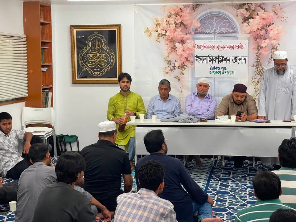 Men seated on a patterned carpet listen to speakers seated behind a long table in front of a banner with Bengali text reading "Islamic Mission Japan," decorated with pink floral fabric and a framed Arabic calligraphy piece on the wall.