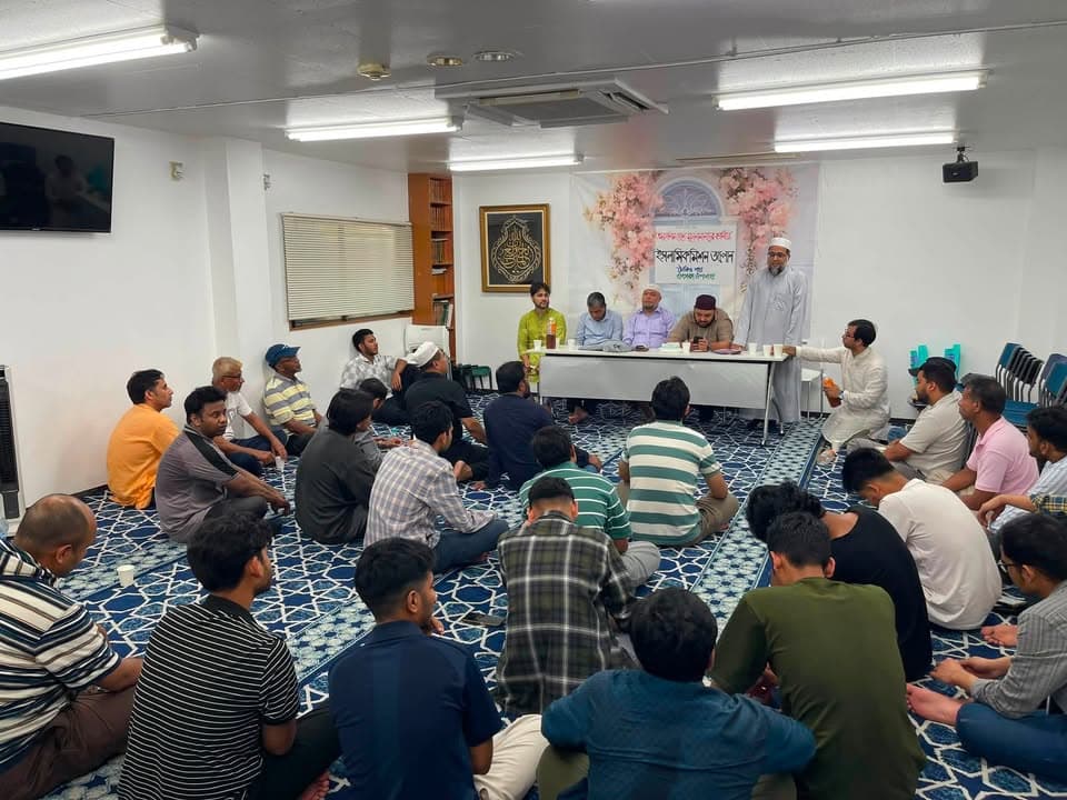 A group of men sit cross-legged on a patterned carpet inside a prayer hall, listening to several speakers seated behind a long table at the front beneath a decorated banner.