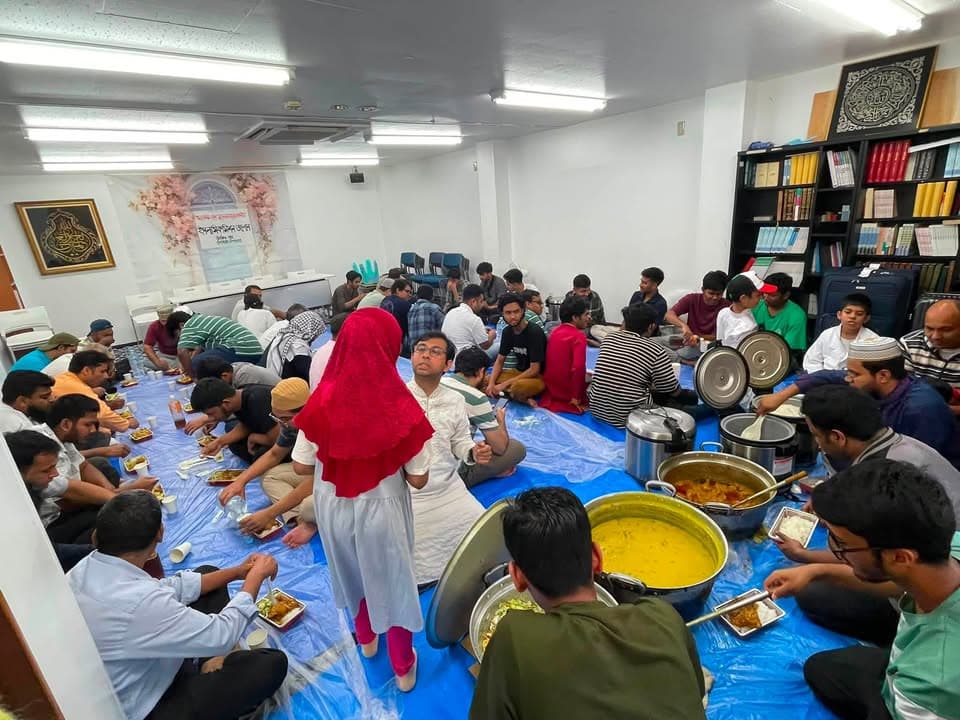 A large group of men and women sit on plastic sheeting on the floor of a room lined with bookshelves, eating a shared meal from trays while large pots of curry and rice sit in the center.