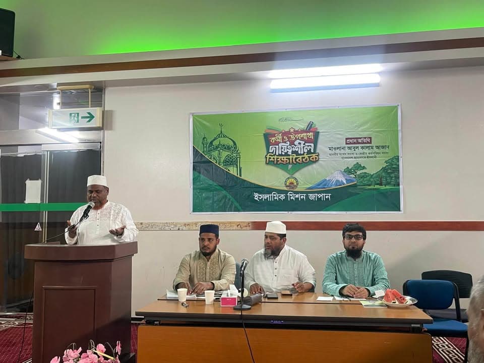 A man in a white kurta and cap speaks at a podium with a microphone, while three seated men in traditional attire listen at a table in front of a green banner with Bengali text reading "Islamic Mission Japan".