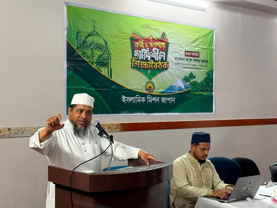A man in white traditional attire and a white cap speaks passionately at a podium with a microphone, gesturing with his hand raised, in front of a large green banner with Bengali text reading "Islamic Mission Japan," while another man in beige attire and a blue cap sits nearby typing on a laptop.