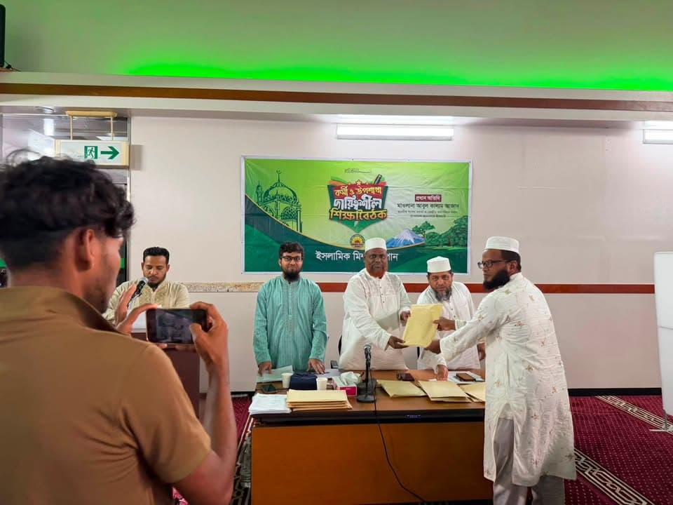 Men in traditional white attire stand behind a table with documents, handing a certificate to another man, in front of a green event banner with Bengali text, while a man in the foreground films the moment on his phone.