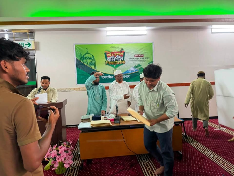 Men gather in a hall decorated with a green banner in Bengali, standing and seated at a table on a red patterned carpet during an indoor gathering.