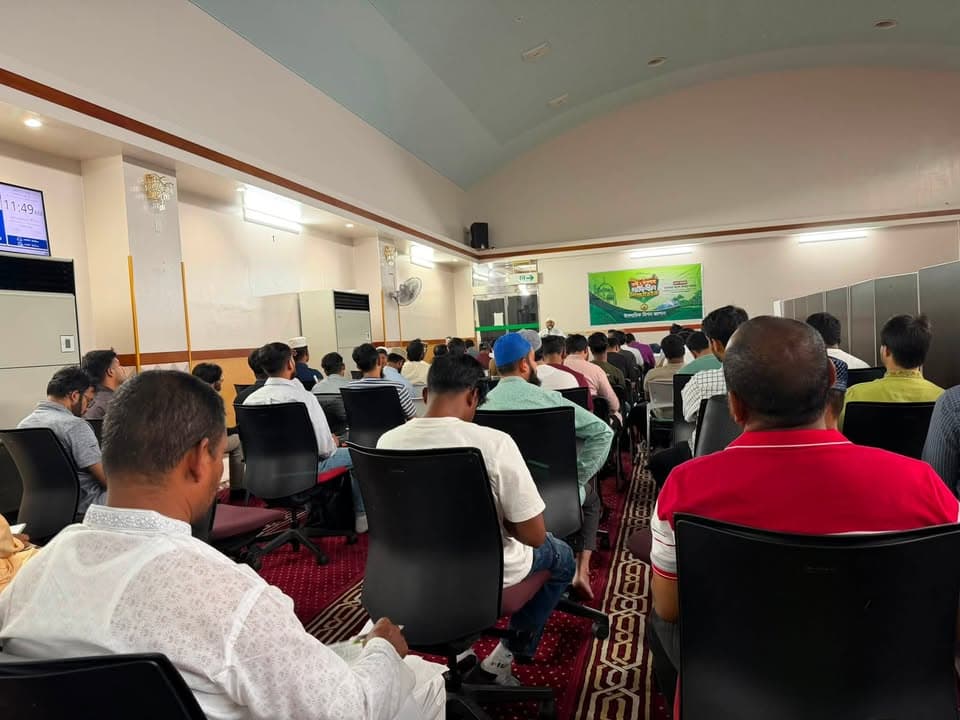 A large group of men sit on chairs in rows inside a hall, facing a speaker at the front near a green banner with text, listening attentively during an indoor gathering.