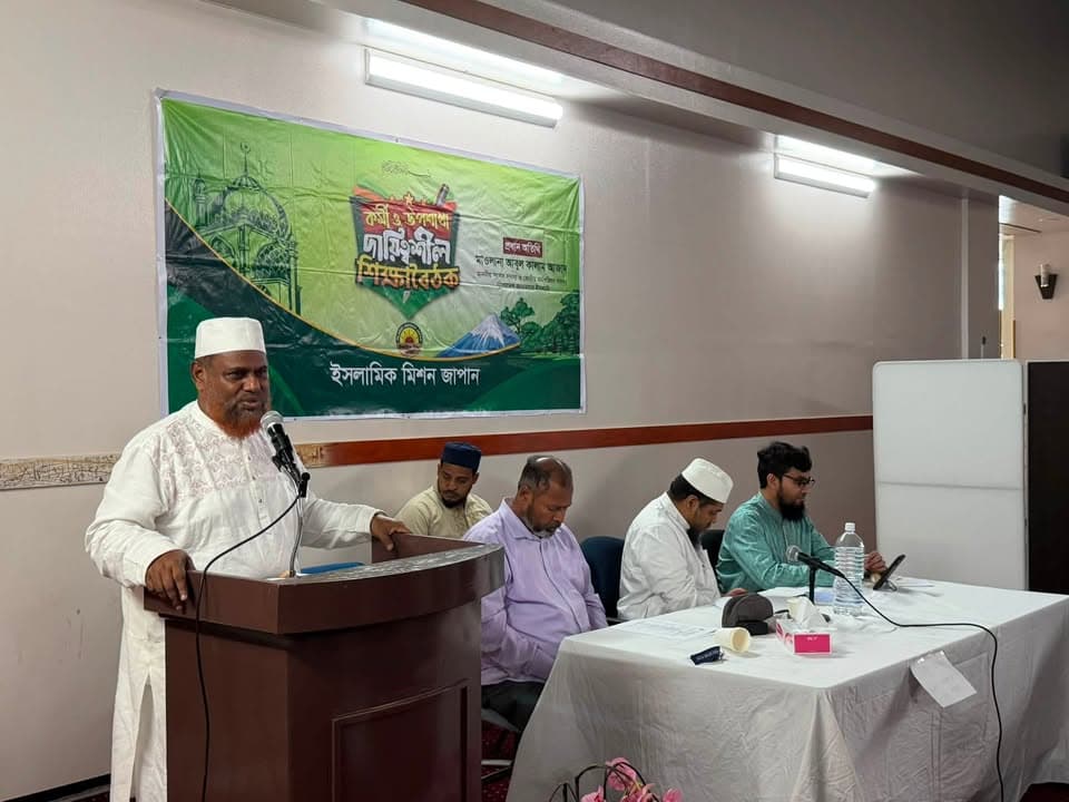 A man in white traditional clothing and a white cap speaks into a microphone at a wooden podium, while four seated men listen at a table covered with a white cloth, in front of a green banner with Bengali and Japanese text reading "Islamic Mission Japan".