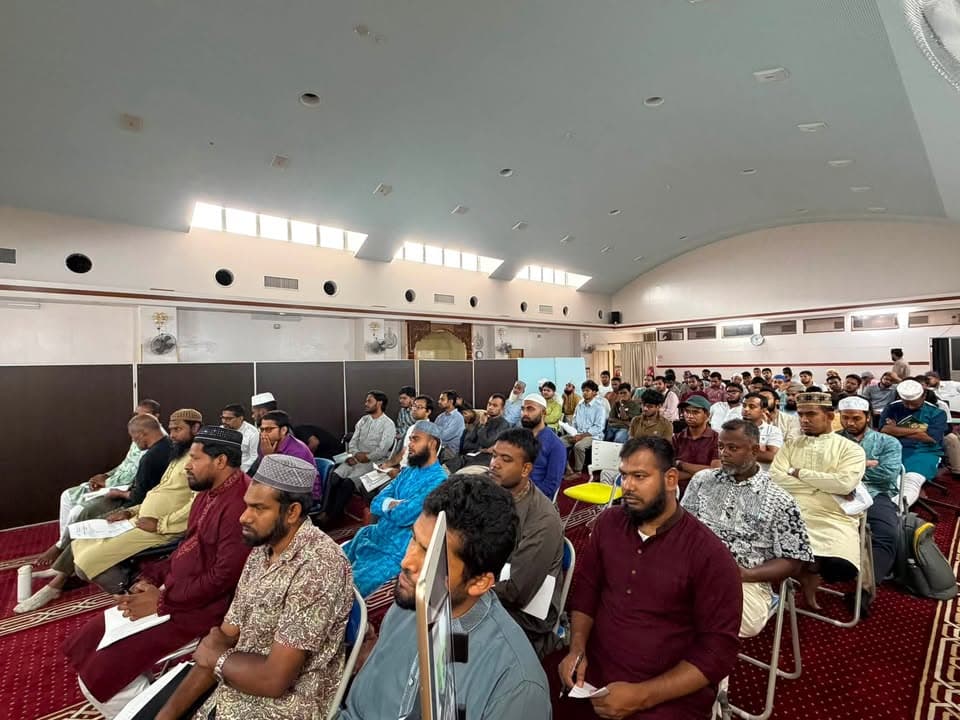 A large group of men sit on chairs and on the carpeted floor inside a hall with a domed ceiling, listening attentively to a speaker at the front, with a projector screen visible in the background.
