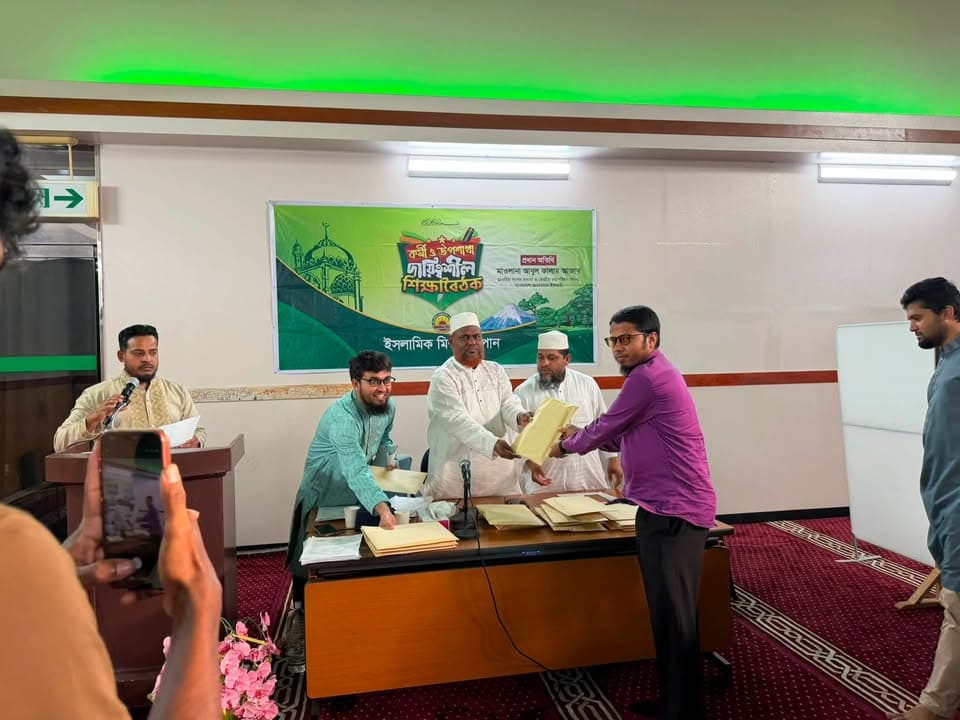 A man in a purple shirt receives a certificate document from seated organizers at a table, in front of a green banner with Bengali text, while a man speaks at a podium and another films the moment on a phone.