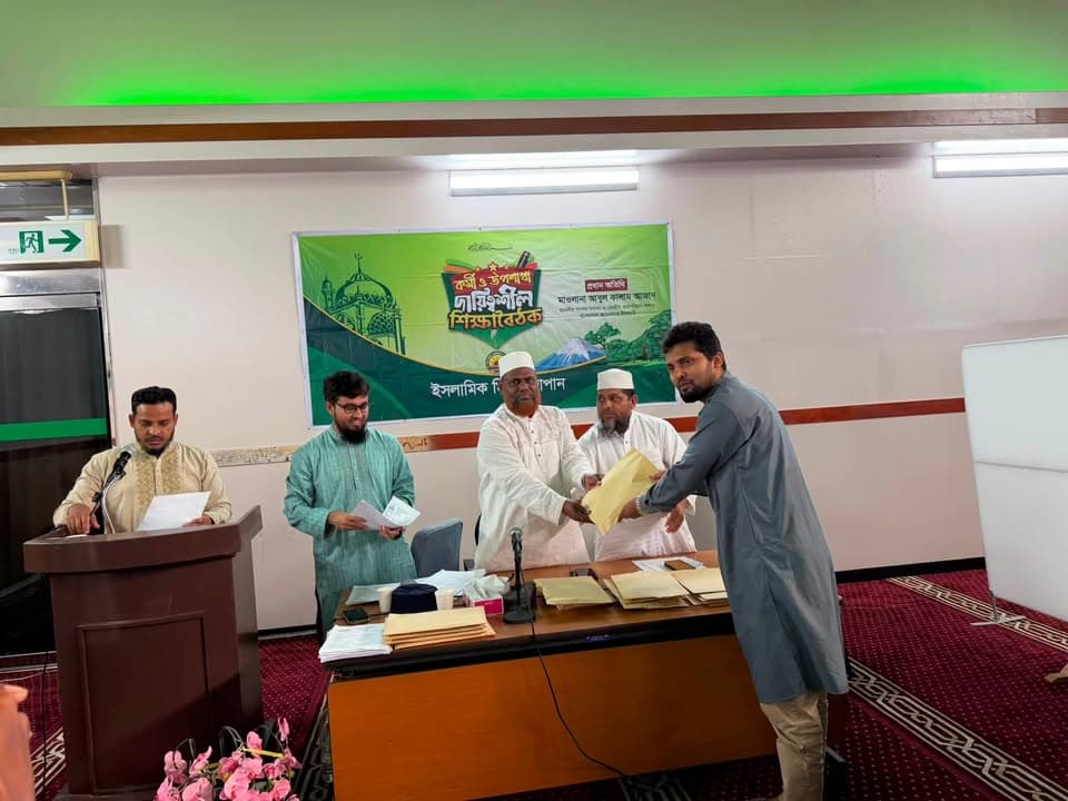 Men in traditional attire stand at a table on a stage, with one handing over a document to another, in front of a green banner with Bengali text reading "Islamic Mission Japan" while a man at a podium reads from papers.