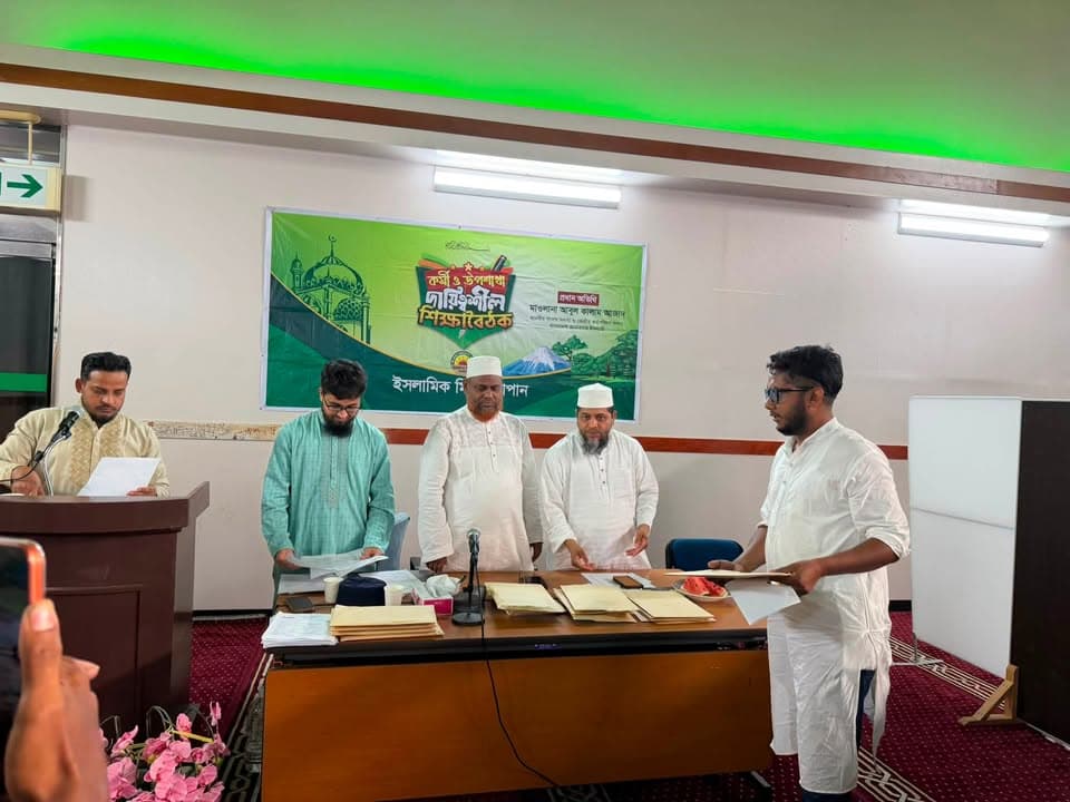 Five men in traditional South Asian attire stand behind a table with documents, one reading from a paper at a podium, in front of a green banner with Bengali text, during an indoor gathering.