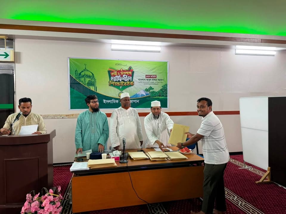Several men in traditional attire stand behind a table covered with envelopes and documents in a hall decorated with a green Bengali-language banner, as one hands an envelope to a participant.