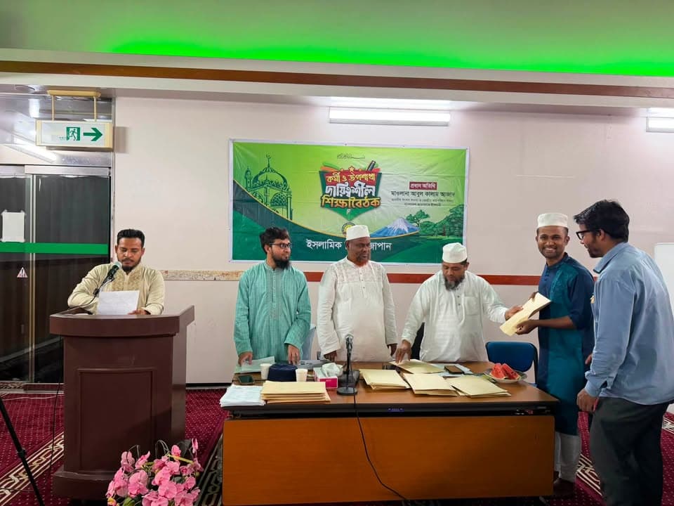 Men in traditional Islamic attire stand behind a table at the front of a hall with a green Bengali-language banner on the wall, one man handing a document to another while a man at a podium reads from a paper.