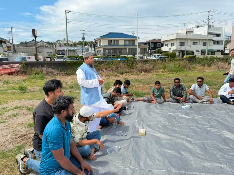 A man in traditional dress stands and speaks while a group of men sit around him on a gray tarp spread on grass, with houses and parked cars visible in the background.