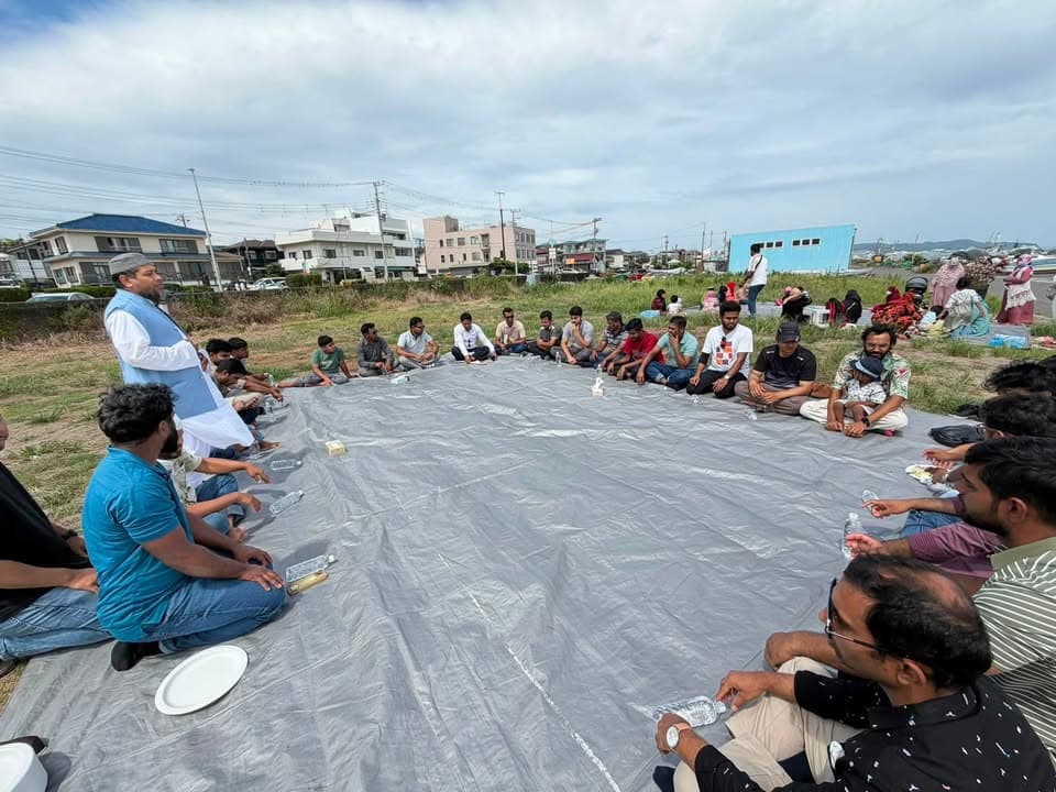 A group of men sit in a large circle on a gray tarp spread over grass near the coast, with one man standing and speaking to the group, water bottles placed in front of the seated attendees, and buildings and the sea visible in the background.